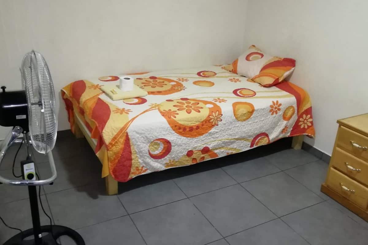 A bedroom with a bed, a standing fan, and a wooden dresser.
