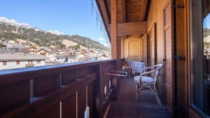 Un balcon en bois, agrémenté de chaises et d’une table en osier, qui offre une vue sur un village avec ses maisons et ses montagnes.
