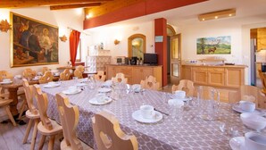 Un coin repas avec des chaises et des tables en bois, un tableau accroché au mur et une cuisine visible à travers une porte ouverte.