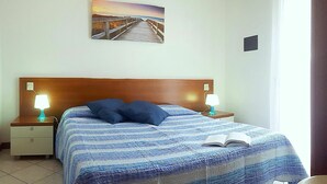 A hotel room with a wooden headboard, a bed with a blue striped bedspread, two bedside lamps, a nightstand, a small table, and a painting of a sunset over a pier. - Nice apartment for 5 guests with WIFI, A/C, TV, terrace and pets allowed (San Michele Al Tagliamento)