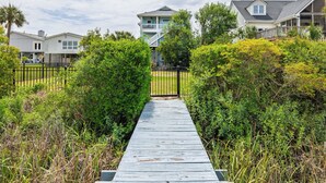 A wooden pathway leading to a two-story house with a balcony, surrounded by lush greenery.