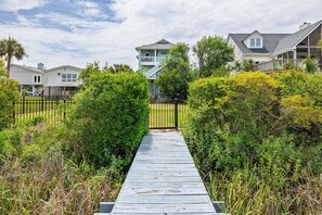 A wooden pathway leading to a two-story house with a balcony, surrounded by lush greenery.