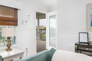 A room with a bed, a bedside table with a lamp, a window with blinds, a door leading to a balcony, and a framed map on a side table.