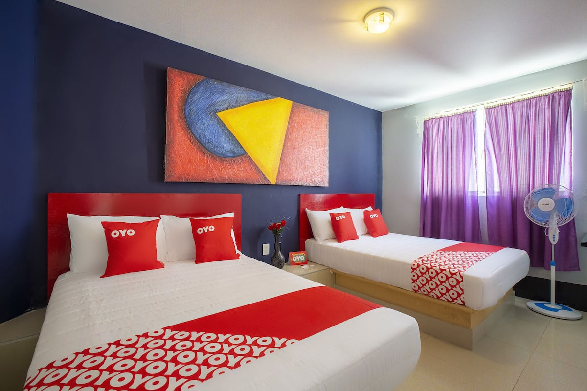 A hotel room with two beds, each with red and white bedding and pillows, a large abstract painting on the wall, and a window with purple curtains.