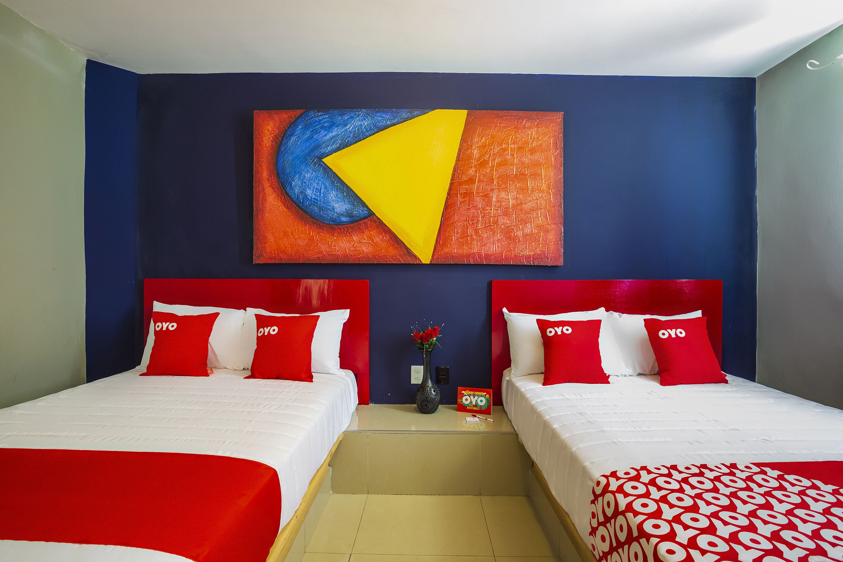 Une chambre d’hôtel avec deux lits, une literie rouge et blanche, un grand tableau abstrait accroché au mur et un vase rempli de fleurs sur la table de chevet.