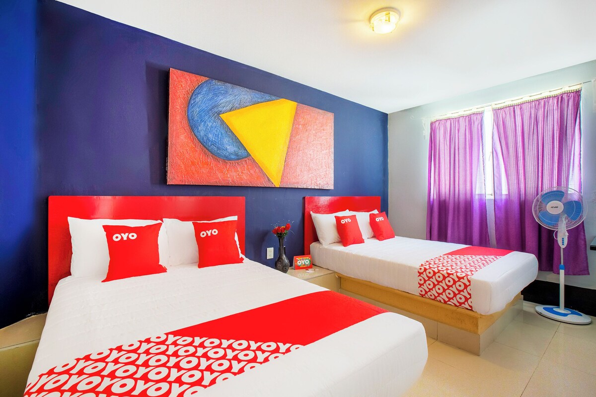 A hotel room with two beds, each with red and white bedding and pillows, a large abstract painting on the wall, and a window with purple curtains.