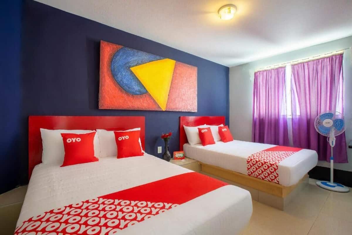A hotel room with two beds, each with red and white bedding, a large abstract painting on the wall, and a window with purple curtains.