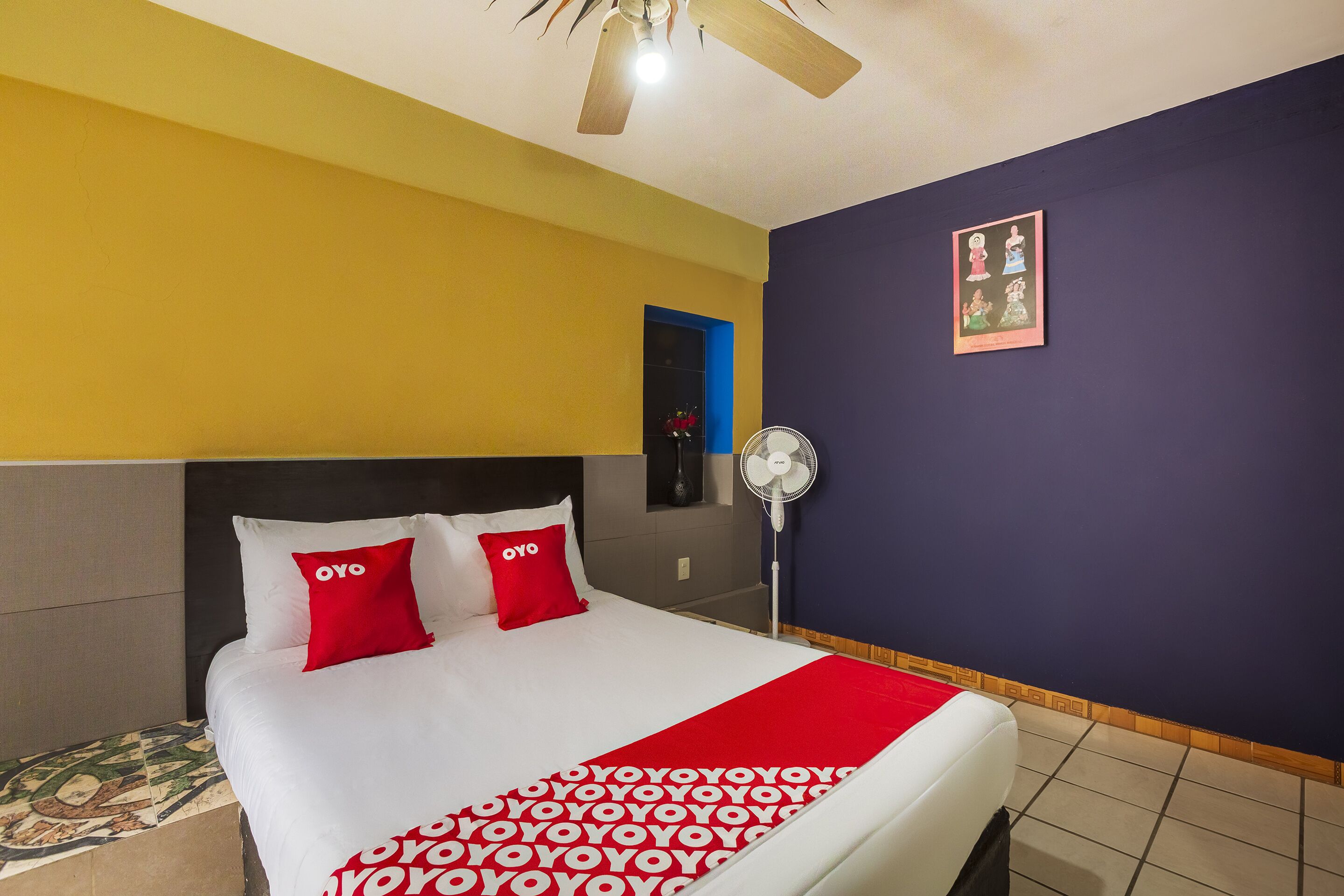 Habitación de hotel con cama, ventilador de techo, ventilador de pared y un cuadro en la pared.