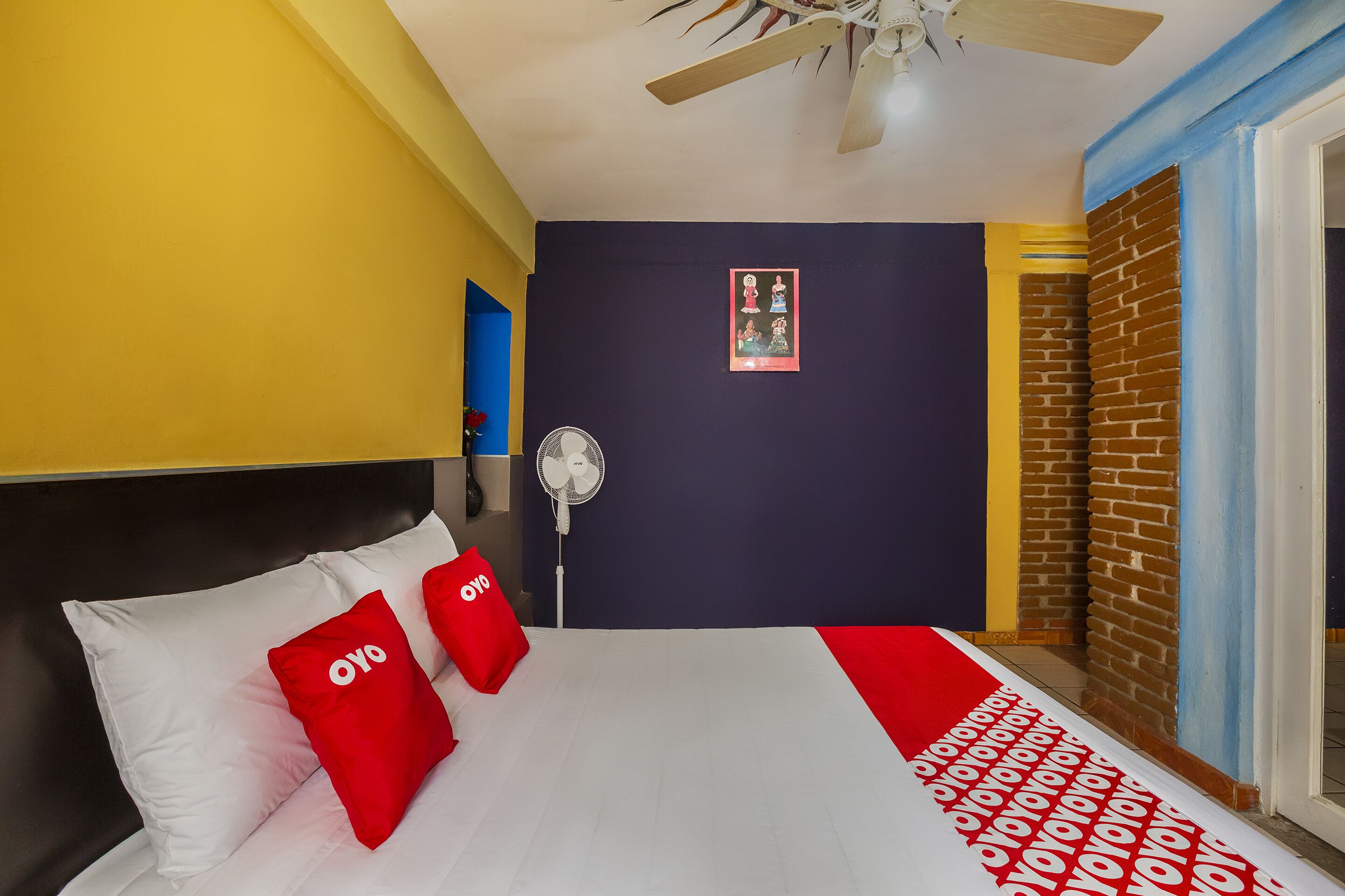 A hotel room with a bed, a ceiling fan, a wall-mounted fan, and a door leading to a balcony.