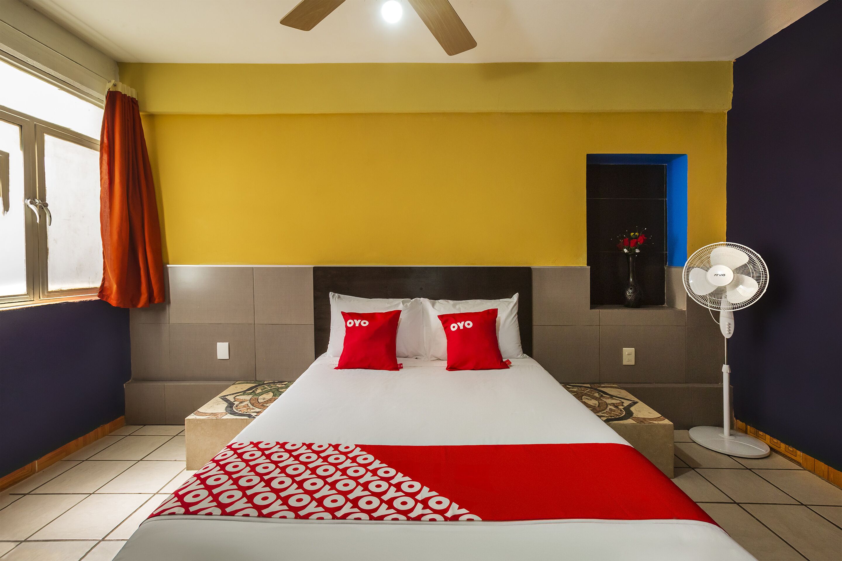 A hotel room with a bed, a standing fan, and a window with curtains.