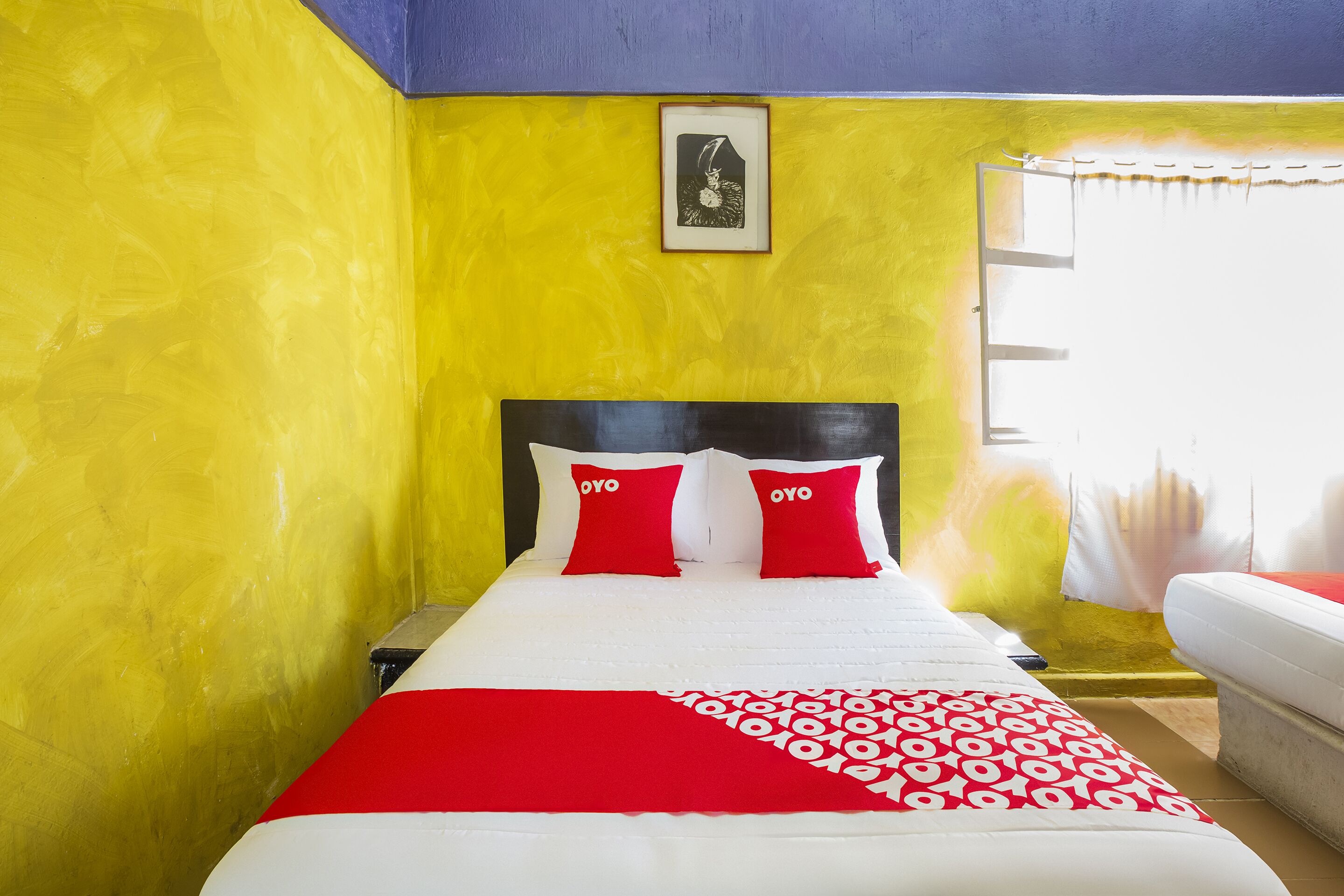 Una cama con ropa de cama roja y blanca, un cuadro enmarcado en la pared y una ventana con cortinas.