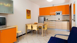 Een kleine keuken met oranje kastjes, een magnetron en een koelkast. Er is een eethoek met een tafel en stoelen, en een televisie op een dressoir.