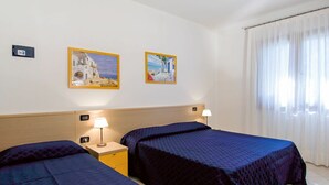 Hotelkamer met twee bedden, een nachtkastje met een lamp, een raam met dunne gordijnen en twee ingelijste foto's aan de muur.