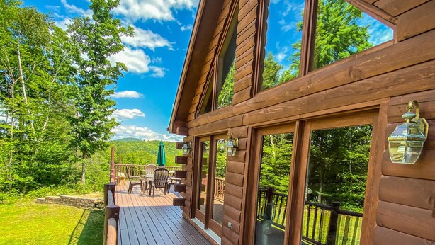 A wooden cabin with a deck, large windows, and a view of the surrounding forest.