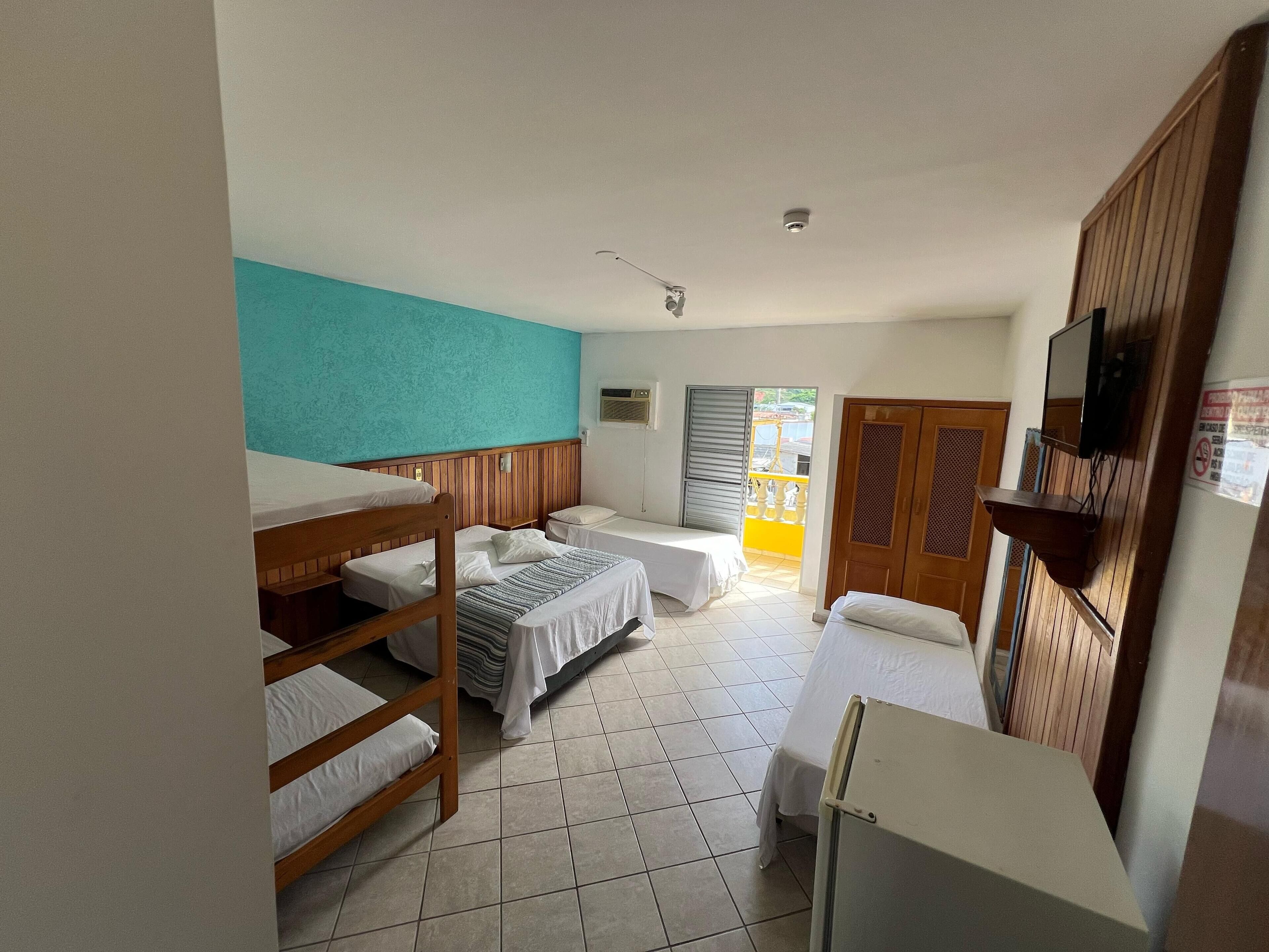 A dormitory room with bunk beds, a small table, a TV mounted on the wall, and a balcony with yellow railings.