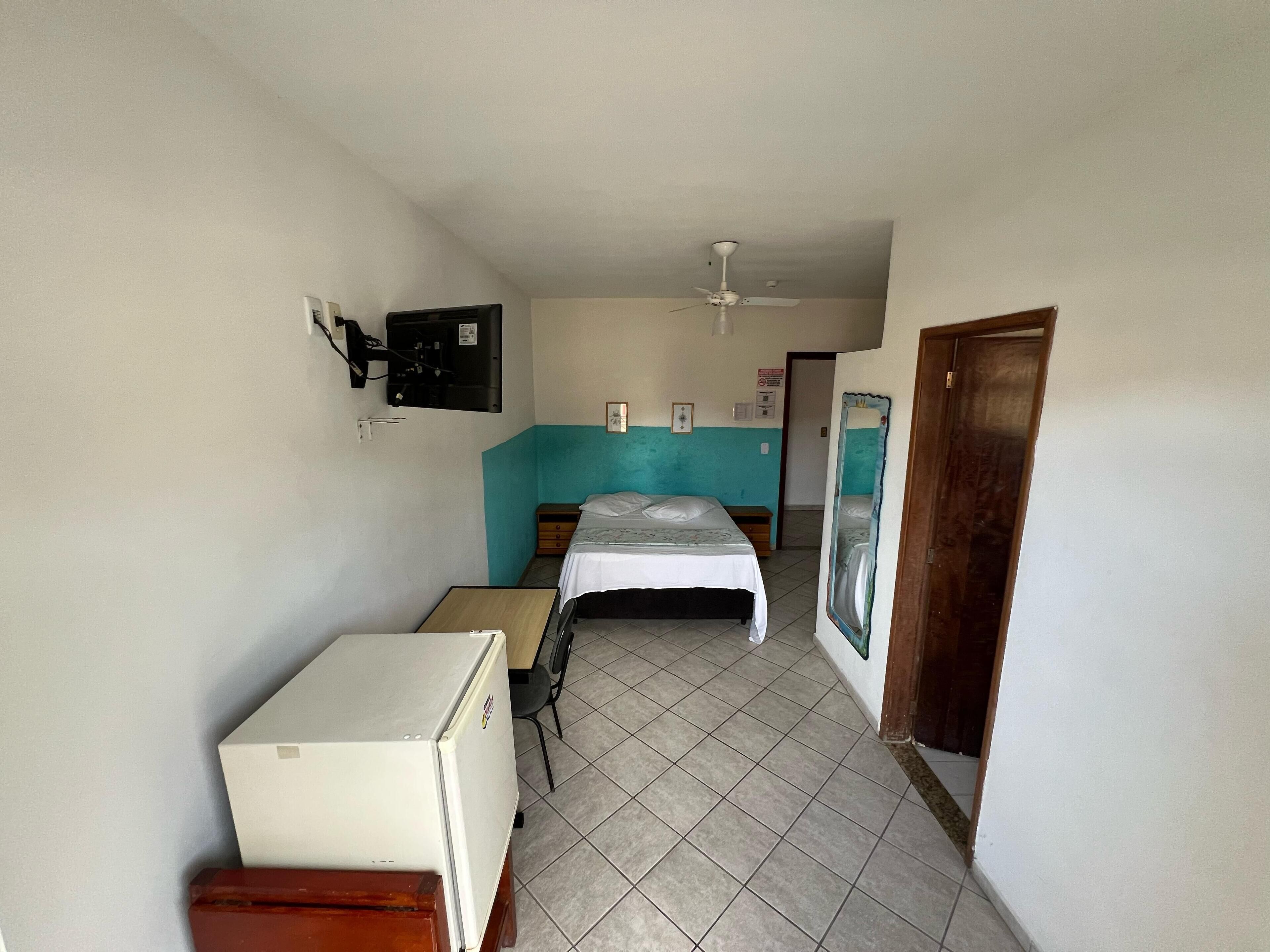 Una habitación pequeña y con pocos muebles, con cama, escritorio, refrigerador y ventilador de techo.