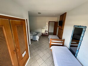 A hotel room with two single beds, a wooden wardrobe, a small desk with a chair, a television mounted on the wall, and a tiled floor. - Pousada Martin de Sá (Caraguatatuba)