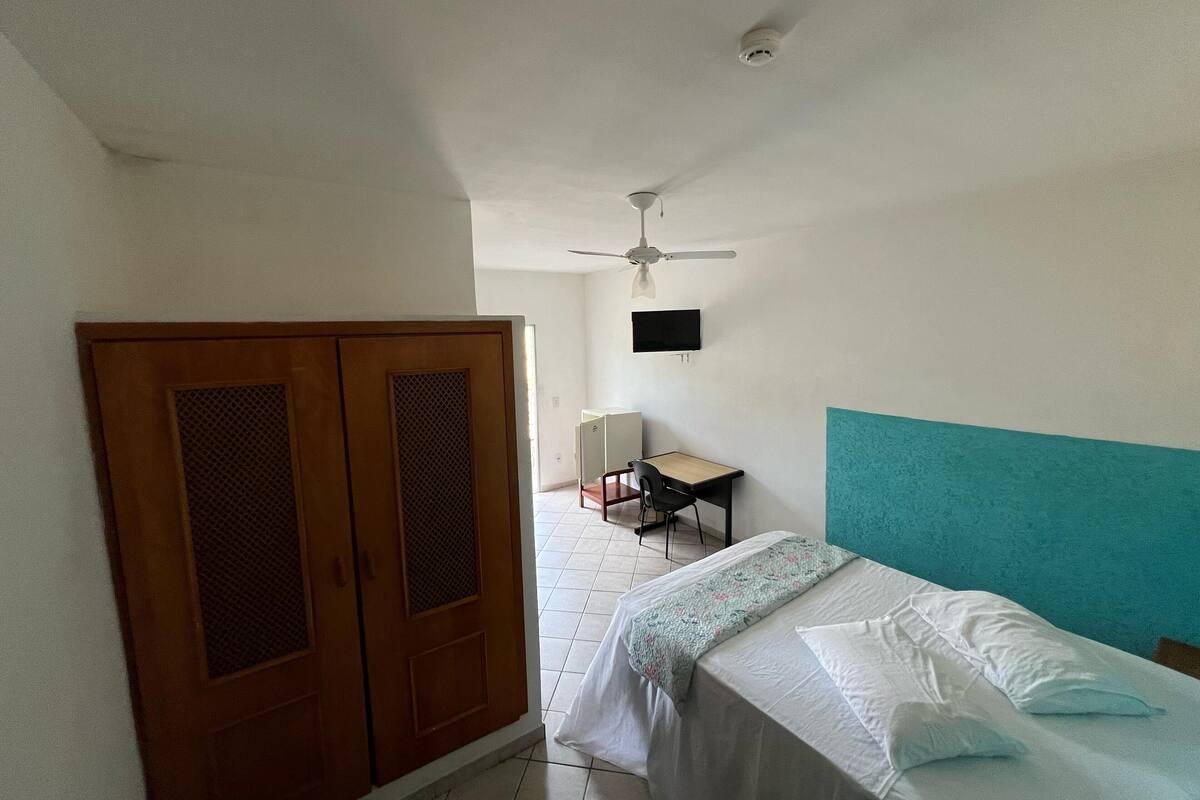 Um quarto com cama, escrivaninha, cadeira, televisão e guarda-roupa.