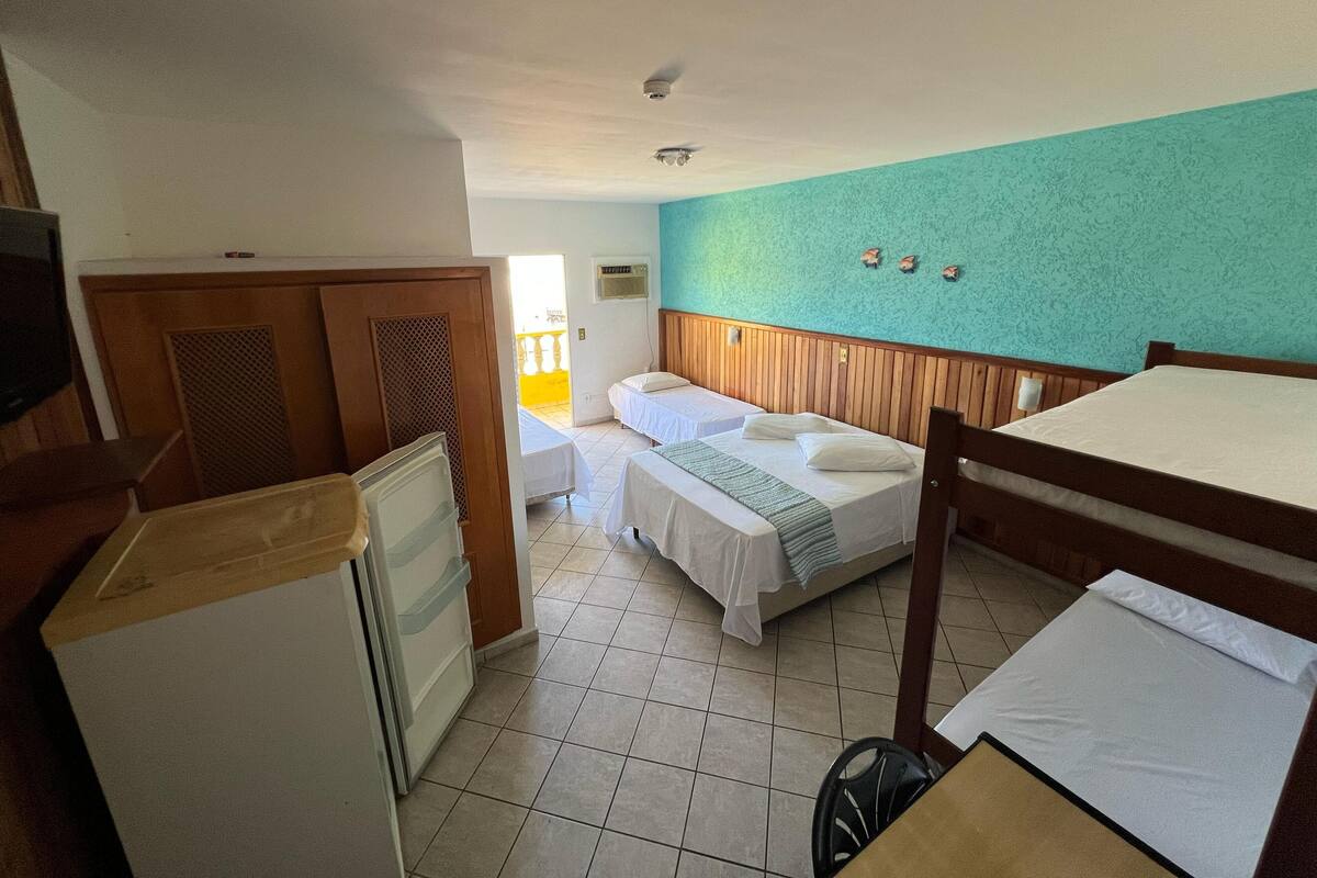 Quarto de hotel com duas camas, um guarda-roupa de madeira, uma pequena cozinha e um banheiro visível por uma porta aberta.