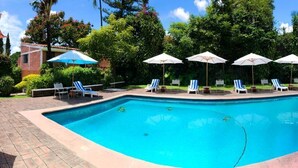 Une piscine rectangulaire à l’eau bleue, entourée de chaises longues et de parasols, avec en arrière-plan une végétation luxuriante et un bâtiment en briques.