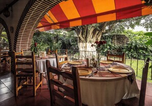 An outdoor dining area with tables set for two, surrounded by a brick wall and a large tree providing shade.