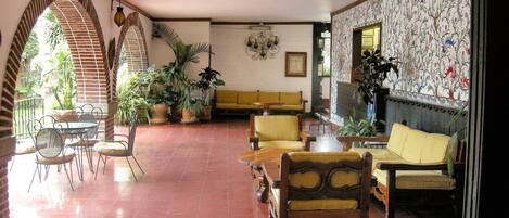 Un espacio interior amplio con suelo de baldosas, un sofá amarillo, varias sillas y plantas en macetas.