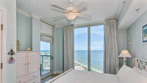 A bedroom with a bed, a ceiling fan, a nightstand, a lamp, a dresser, and a view of the ocean. - Summer is here @ Portofino 2-1409! (Pensacola Beach)