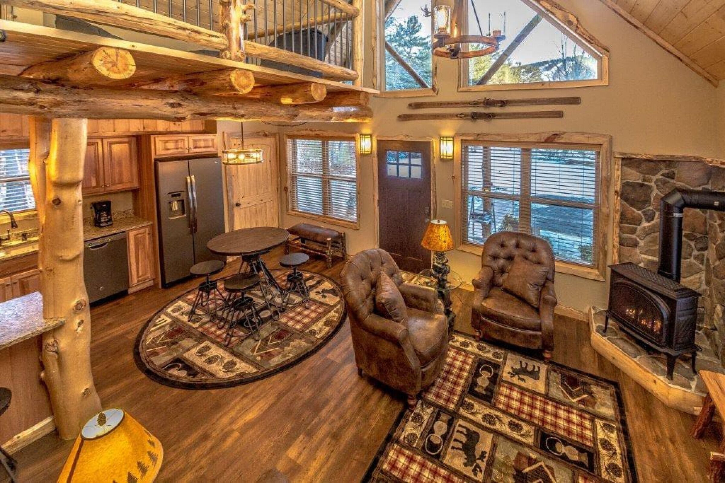 A rustic cabin interior with a wooden dining table, two leather armchairs, a stone fireplace, and a large window overlooking a forest.