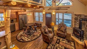 A rustic cabin interior with a wooden dining table, two leather armchairs, a stone fireplace, and a large window overlooking a forest.