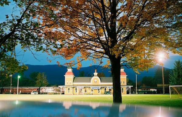 A building with a large tree in front, a reflection in the water, and mountains in the background.