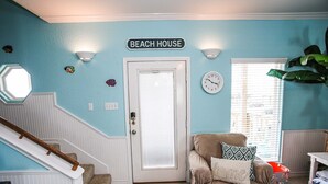 Una habitación con una escalera, una puerta con la inscripción "BEACH HOUSE", un reloj y una ventana con persianas.
