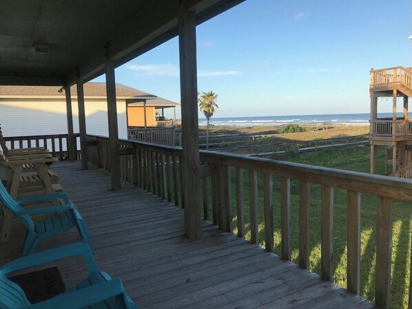 Eine Holzterrasse mit Geländer, die auf einen Strand blickt, mit Stühlen und einem überdachten Bereich im Hintergrund.