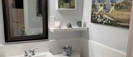 A bathroom with a white sink, toilet, and mirror. There is a shelf with a small chair and a decorative item.