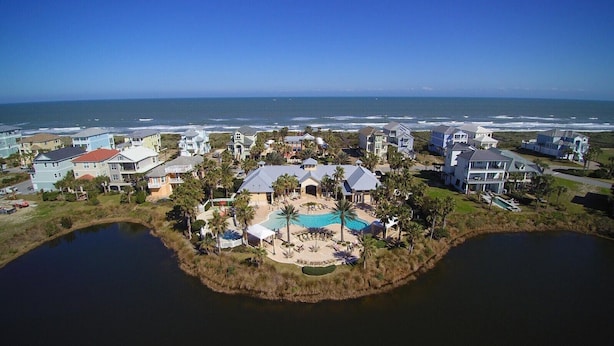 An aerial view of a coastal residential area with houses, a central swimming pool, and a small island with a dock.