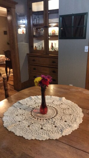 Une table à manger recouverte d’une nappe au crochet, un vase rempli de fleurs colorées et, au fond, un buffet en bois doté de portes en verre.