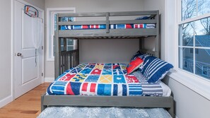 Un lit superposé avec une couverture aux couleurs vives, une fenêtre donnant sur un beau paysage et une porte surmontée d’un panier de basket.