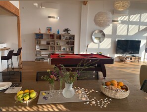 A modern living room with a pool table, a bookshelf, a dining area with chairs, and a TV.