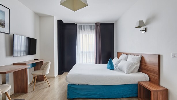 A modern hotel room with a bed, bedside table, desk, chair, and a television. - Chambre Confortable et Abordable près du RER (Rueil Malmaison)