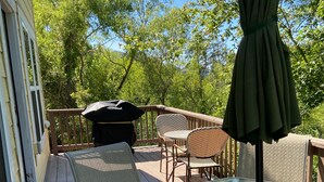 A covered outdoor patio with a glass table and chairs, surrounded by a wooden deck and a view of trees. - Cozy 3br cottage on sound side near beach (Kill Devil Hills)