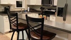 A kitchen with white cabinets, a stainless steel refrigerator, a microwave, and two bar stools. - Cozy 3br cottage on sound side near beach (Kill Devil Hills)