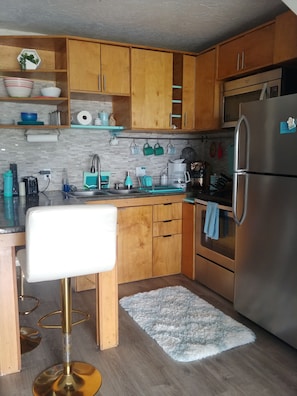 A compact kitchen with wooden cabinets, a stainless steel refrigerator, and a bar stool. - Full Ocean View! Prime Kihei Location!  (Kihei)