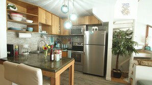 A compact kitchen with wooden cabinets, stainless steel appliances, a dining area with a table and chairs, and a potted plant. - Full Ocean View! Prime Kihei Location!  (Kihei)