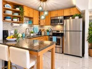 A modern kitchen with wooden cabinets, stainless steel appliances, a marble countertop, and bar stools. - Stunning Upstairs Unit with Direct Ocean View, Steps to  Beach.  Heart of Kihei (Kihei)