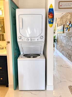 A front-loading washing machine in a hallway with a teal door, a tiled floor, and a wall with a surfboard and a framed quote. - Stunning Upstairs Unit with Direct Ocean View, Steps to  Beach.  Heart of Kihei (Kihei)