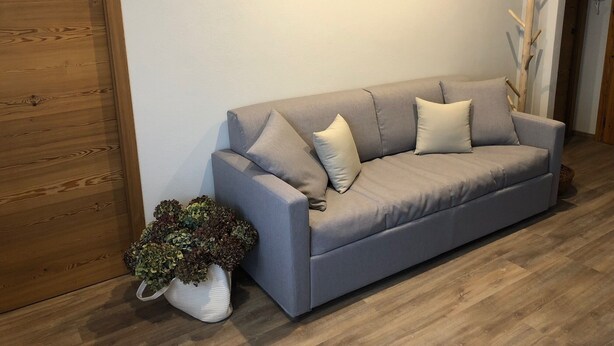 En grå sofa med puder, en hvid vase med en plante, og et trægulv.