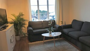 Ein Wohnzimmer mit zwei grauen Sofas, einem weißen Couchtisch, einem Fernseher auf einem Schrank und einem großen Fenster mit Vorhängen.