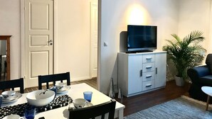 Ein Wohnzimmer mit Fernseher auf einem Schrank, einem Esstisch gedeckt mit Schüsseln und Tassen, einer Couch und einer Topfpflanze.