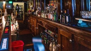 Un bar avec des boiseries, un téléviseur fixé au-dessus du comptoir, et diverses bouteilles et verres disposés sur des étagères.