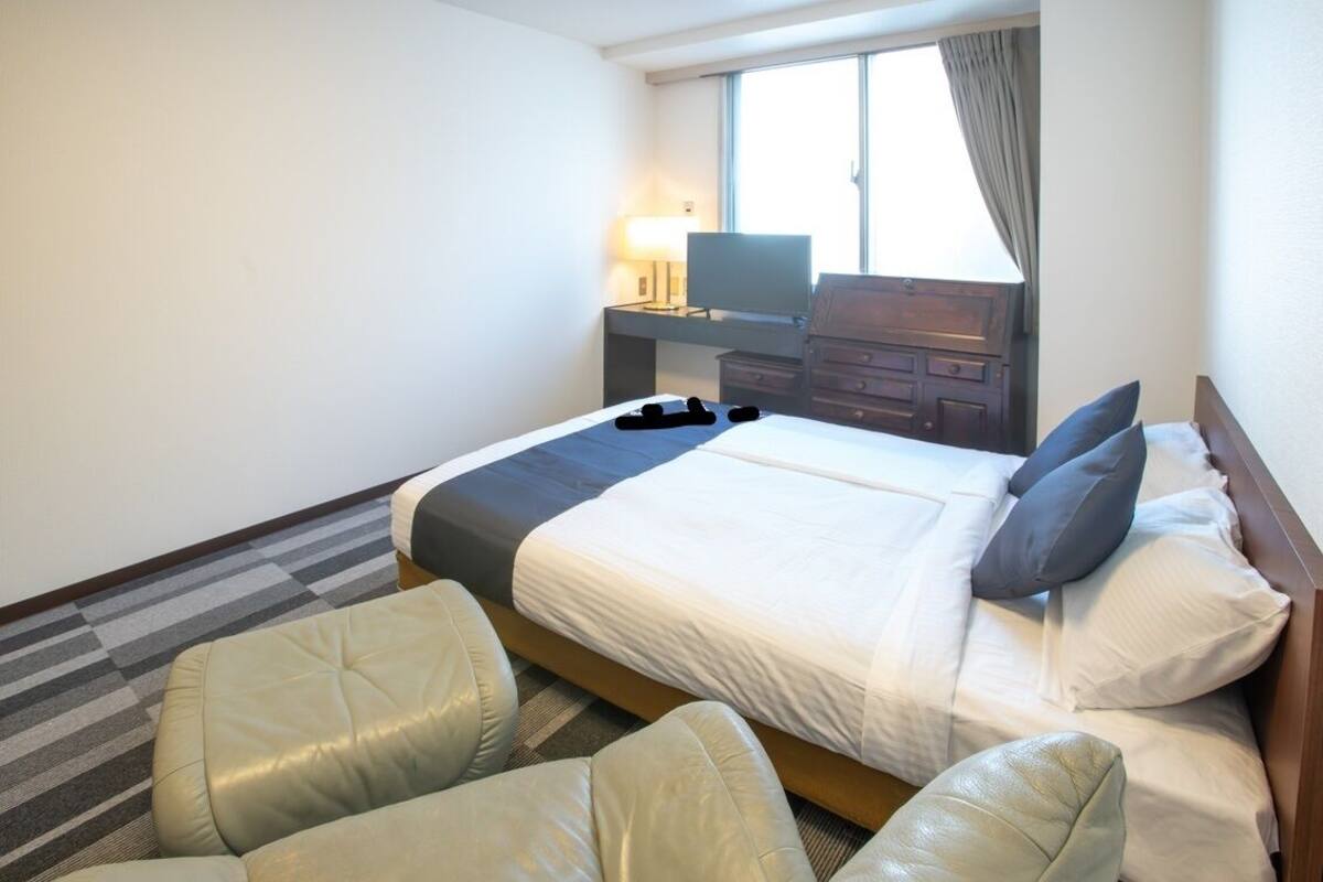 A hotel room with a bed, two chairs, a desk with a lamp, and a TV.