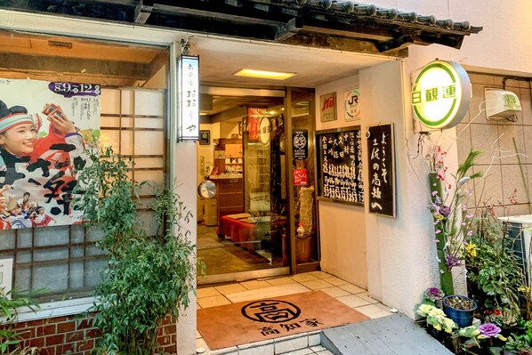 L’entrée d’une auberge japonaise traditionnelle, avec une porte coulissante, un paillasson et une enseigne portant des caractères japonais.